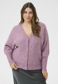 Vrouw met lang bruin haar, gekleed in een zacht roze V-hals trui en een donkergrijze rok, staande tegen een effen lichte achtergrond.
