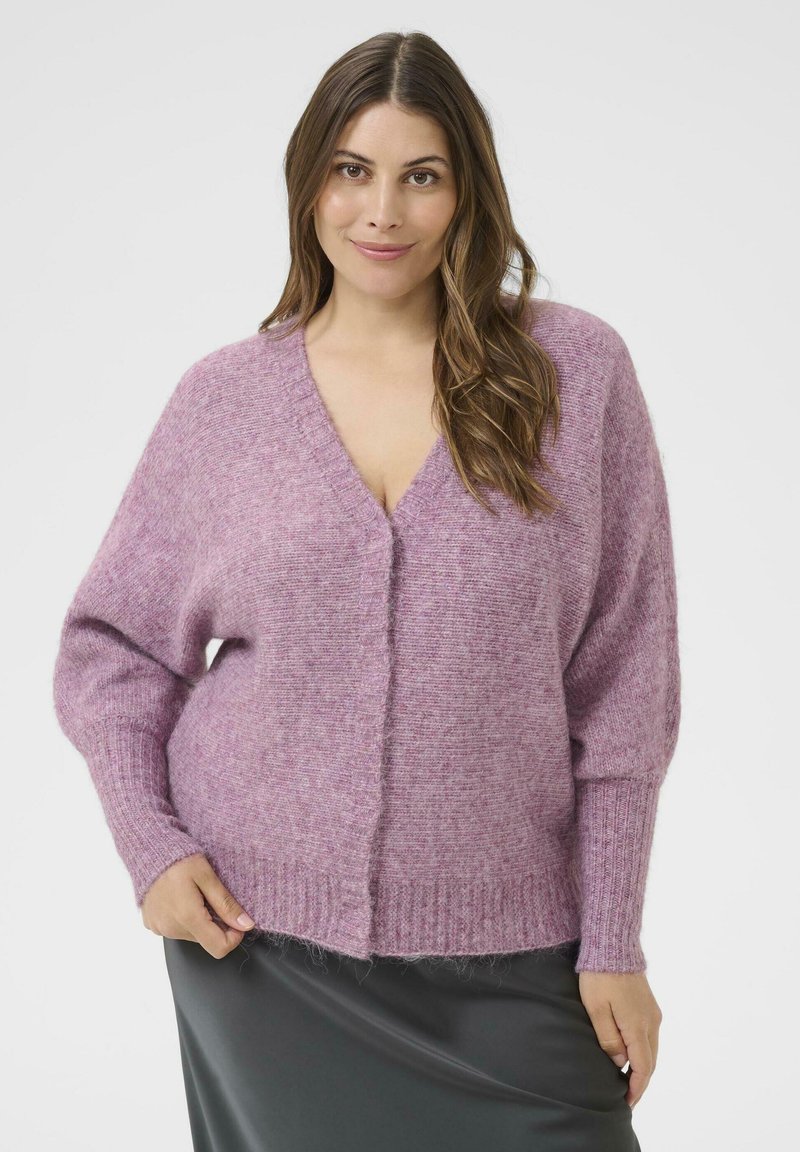 Vrouw met lang bruin haar, gekleed in een zacht roze V-hals trui en een donkergrijze rok, staande tegen een effen lichte achtergrond.