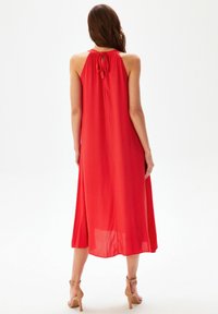 Robe sans manches rouge à coupe fluide, détail de noeud dans le dos, tissu léger et ourlet au niveau des genoux. Texture lisse et couleur unie.