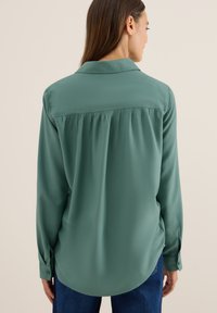 Blouse verte à manches longues en tissu lisse, avec un ourlet incurvé, des plis au dos et des poignets boutonnés.