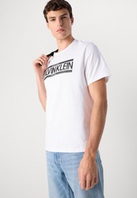 Man wearing white Calvin Klein T-shirt and light blue jeans, holding black strap over shoulder, plain light background.