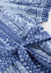 Blått mönstrat tyg har paisleymotiv, blommönster och geometriska former. Det har en knytframsida och knapppdetaljer, med en mjuk textur.