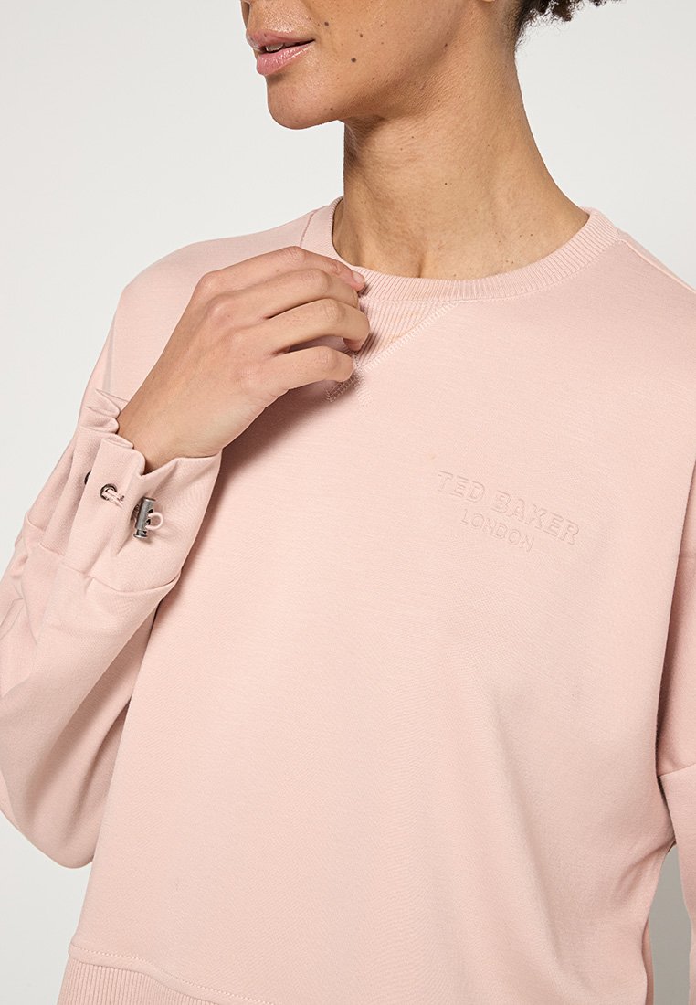 Person, der einen hellrosa Sweatshirt von Ted Baker London trägt, hält den Stoff nahe am Kragen, mit sichtbarem Knopfdetail an der Manschette des Ärmels.