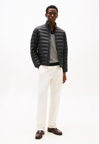 Homme portant une veste matelassée noire, un pull rayé noir et blanc, un pantalon blanc et des mocassins marron foncé, debout devant un fond blanc.