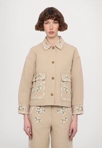 Beige jacket with floral embroidery, featuring a collar, two front pockets, and gold buttons. Smooth fabric with detailed stitching.