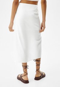Falda blanca, hasta la rodilla, con una textura suave y un diseño sencillo. Combinada con sandalias marrones con cordones que tienen una suela gruesa.