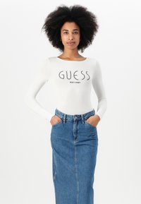 Valkoinen pitkähihaisuus bodysuit, jossa on musta "GUESS"-logo ja "EST.1981"-teksti, yhdistettynä korkeavyötäröiseen farkkuhameeseen.