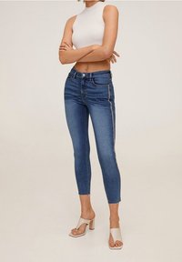 Jeans ajustados de mezclilla azul con una cintura alta, que cuentan con un cierre de botón, cinco bolsillos y bandas laterales en contraste. Combinados con una blusa blanca de canalé y tacones.