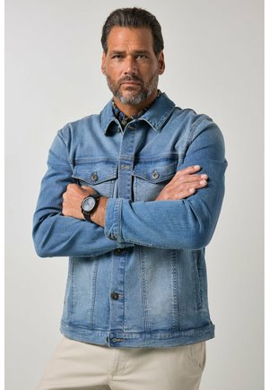 Mann mit Bart, der eine blaue Jeansjacke trägt, verschränkte Arme und eine schwarze Armbanduhr hat, steht vor einem schlichten hellen Hintergrund.