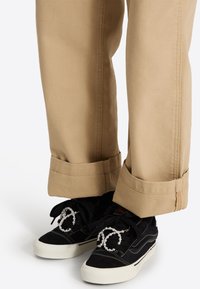 Baskets noires avec semelles blanches et lacets décoratifs imitant des perles, portées avec un pantalon beige à revers.