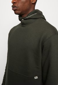 Ciemnozielony sweatshirt z wysokim kołnierzem, teksturowana tkanina oraz metka z napisem "BLEND" na hemie. Subtelny detal w postaci przeszycia na kołnierzu.