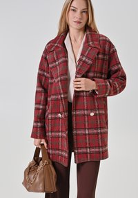 Donna che indossa un cappotto a scacchi rosso e grigio sopra una camicia bianca, mentre tiene una borsa in pelle marrone con una catena dorata su uno sfondo bianco.