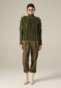 Maglione a collo alto lavorato a maglia verde con dettagli frangiati, abbinato a pantaloni a pieghe verde oliva e stivaletti crema.