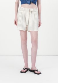 Shorts crème rayés avec une taille élastique, un détail de nouage à l'avant et un tissu léger. Associés à des tongs noires et un design minimaliste.