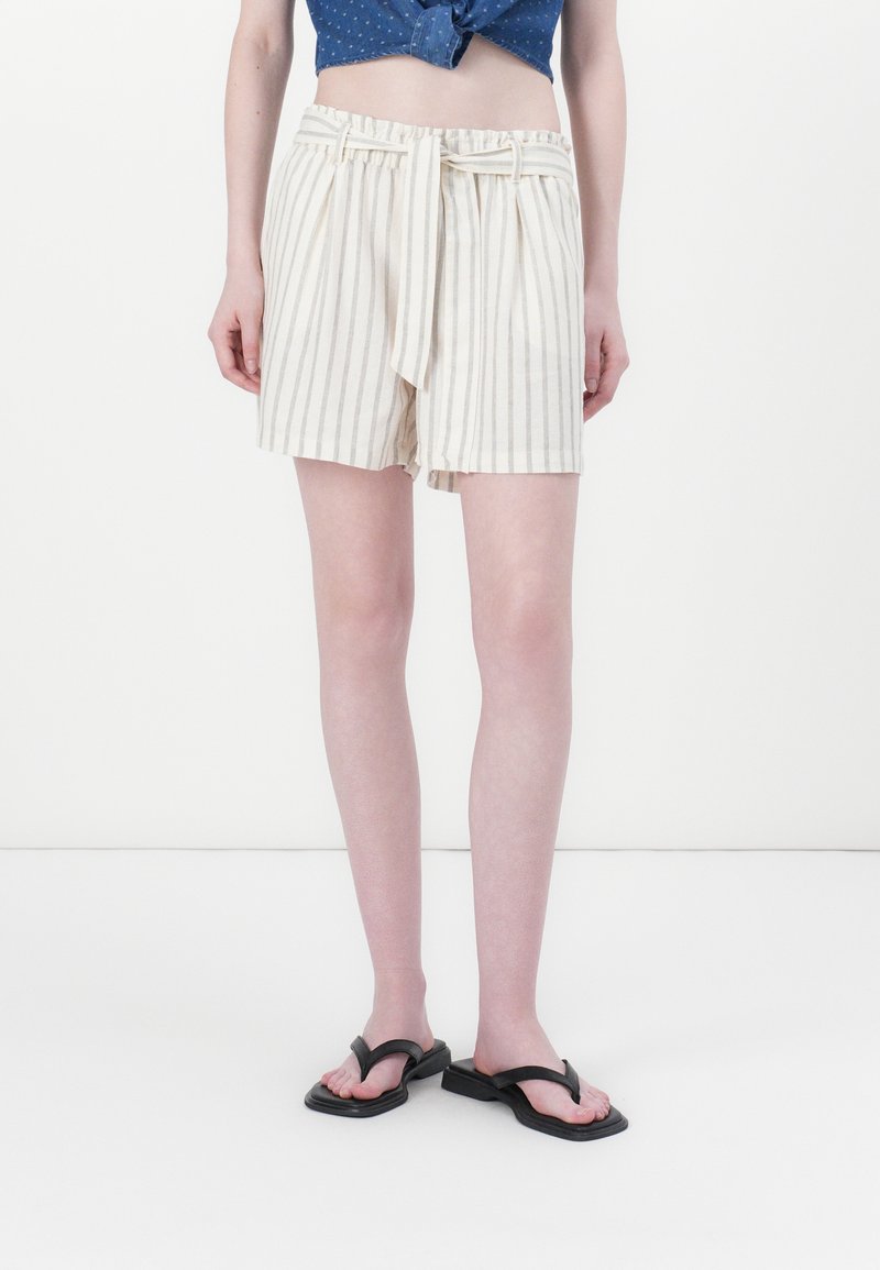Shorts crème rayés avec une taille élastique, un détail de nouage à l'avant et un tissu léger. Associés à des tongs noires et un design minimaliste.