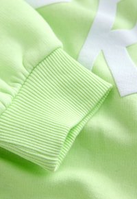 Világos zöld pulóveranyag bordázott mandzsetta textúrával. Nagyméretű fehér grafikai dizájnt tartalmaz, puha, sima felülettel.