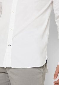 Camicia bianca in cotone a maniche lunghe con chiusura a bottoni, piccolo dettaglio ricamato vicino all'orlo e orlo inferiore curvo.