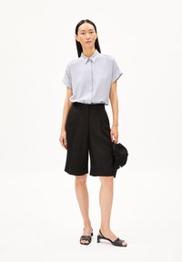 Lichtgrijze korteklevenshirt met kraag en zwarte geplooide Bermudashorts, gecombineerd met zwarte hoge sandaaltjes en een donkere pet.