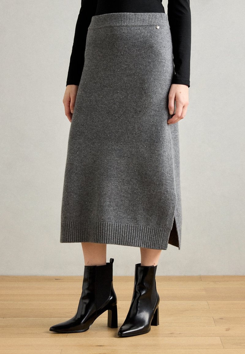 Femme portant une jupe midi en tricot gris avec fente latérale et bottines à talons noires à bout pointu sur un sol en bois clair.