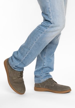 Person trägt hellblaue Jeans und braune Schnürschuhe mit Gummisohlen, steht mit einem leicht angehobenen Fuß.