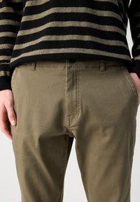 Pantaloni verde măsliniu cu un finisaj neted și texturat, având un buzunar frontal și o cusătură subtilă. Asortați cu un pulover în dungi negre.