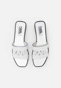 Sandali bianchi con logo ritagliato, suola piatta e bordi neri. Fabbricati in Italia, presentano un materiale liscio e una forma rettangolare.
