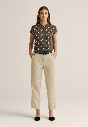 Nainen seisoo kädet taskuissa, hänellä on päällään ruskea pilkullinen paita, beiget housut, musta vyö ja mustat ballerinat yksiväristä taustaa vasten.