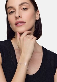 Srebrna bransoletka z zawieszkami w kształcie gwiazdek, przymocowana do smukłej łańcuszka, noszona na ręce spoczywającej na brodzie, w zestawie z czarnym swetrem.