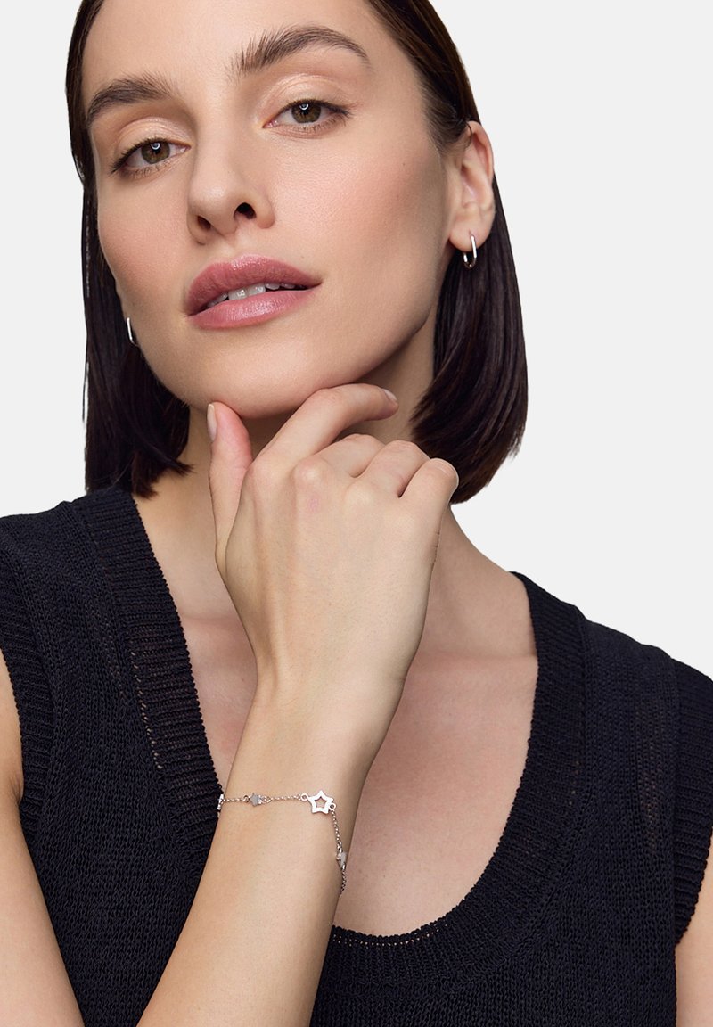 Srebrna bransoletka z zawieszkami w kształcie gwiazdek, przymocowana do smukłej łańcuszka, noszona na ręce spoczywającej na brodzie, w zestawie z czarnym swetrem.