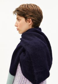 Personne avec les cheveux bruns courts portant un pull en tricot torsadé violet clair et une grande écharpe en laine bleu foncé sur fond blanc.