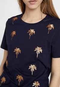 Námořnická modrá bavlněná tričko s rozptýlenými zlatými a bronzovými flitrovými vzory palmy. Krátké rukávy a kulatý výstřih.