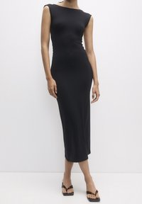 Robe midi noire moulante avec manches courtes, encolure bateau, et texture lisse, complétée par des sandales noires plates à brides.