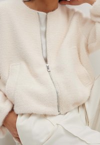 Chaqueta bomber texturizada de color rosa claro con una cremallera plateada, dos bolsillos laterales y un escote redondeado, combinada con una blusa blanca.