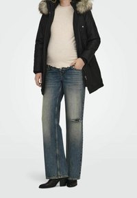 Osoba v černé parce s kožešinovou kapucí, béžovém pleteném svetru, širokých roztrhaných modrých džínách a černých botách na podpatku, stojí s rukama v kapsách.