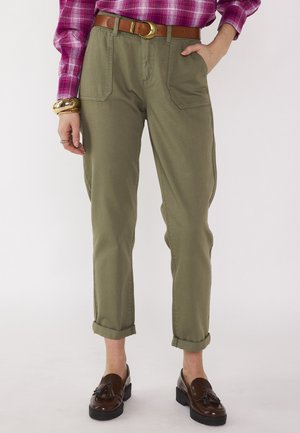Donna con pantaloni verde oliva risvoltati con tasche frontali, cintura marrone, camicia a quadri rosa, braccialetto d'oro e mocassini marroni con nappine.