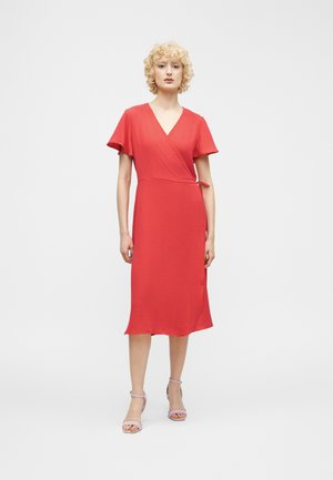 Kobieta o kręconych blond włosach, ubrana w czerwoną sukienkę kopertową do kolan i jasnoróżowe buty na obcasie z odkrytymi palcami, stojąca na jednolitym białym tle.