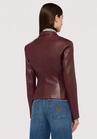 Vínově červená kožená bunda s přiléhavým střihem, límcem a hladkou texturou. Kombinována s modrými džíny s vyšívaným vzorem na kapse.