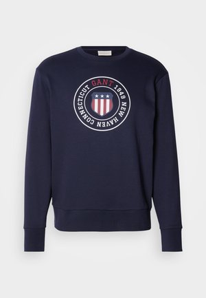 Felpa blu navy in morbido tessuto con una grafica rotonda che presenta uno scudo e stelle, testo "GANT 1949 NEW HAVEN CONNECTICUT" in bianco e rosso.