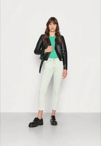 Jaqueta de couro preta, top verde canelado, calças verde-claro e sapatos de plataforma pretos. Modelo está encostado contra um fundo neutro.