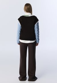 Bruine mouwloze trui over een lichtblauw gebreid overhemd met lange mouwen en een witte overhemdkraag, gecombineerd met een broek met wijde pijpen in bruin.