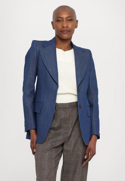 Denim blue blazer with a notched lapel, single-button closure, and two front pockets. Worn over a white knit sweater, paired with plaid trousers.