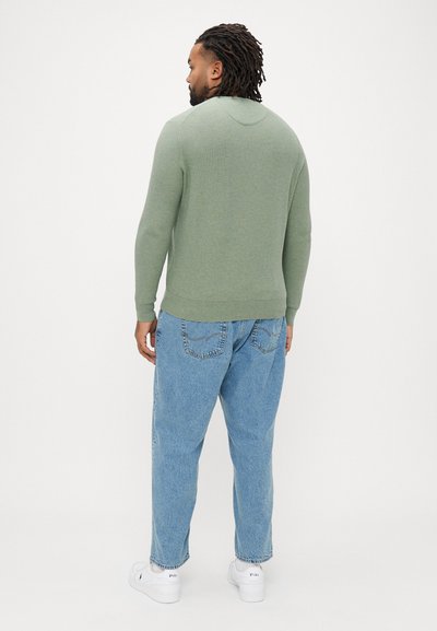 Πράσινο πλεκτό φούτερ με στρογγυλή λαιμόκοψη, ριμπ μανσέτες και τελείωμα, συνδυασμένο με φαρδιά ανοιχτό μπλε τζιν και λευκά αθλητικά παπούτσια.