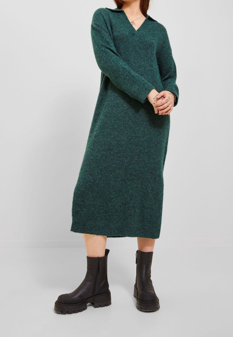 Femme portant une longue robe tricotée vert foncé avec un col et des bottines noires épaisses, debout, les mains jointes.
