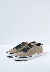 Zapatillas de lona beige con cordones azul marino, suela blanca y acentos rojos y azules. Cuentan con una etiqueta de marca en la lengüeta y una superficie texturizada.