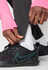 Μαύρα αθλητικά παπούτσια με πράσινες λεπτομέρειες, φορεμένα με λευκές κάλτσες, συνδυασμένα με μαύρο παντελόνι με φερμουάρ και φωτεινό ροζ μπλουζάκι μακρύ μανίκι.