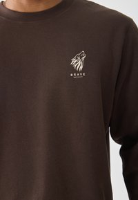Donkerbruine sweatshirt met een witte gestileerde logo van een huilende wolf en de tekst "BRAVE SPIRIT" op de borst.