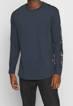 Homme portant une chemise à manches longues bleu marine avec des détails rayés noirs le long des manches et un pantalon noir.