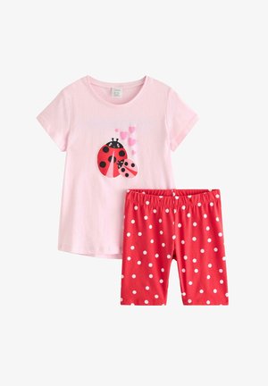 Ung pige med krøllet hår iført en lyserød kasket, lyserød marihøne-t-shirt, røde shorts med prikker og stribede sokker med sneakers.