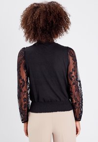 Blouse noire avec un corps en tricot uni et des manches longues en dentelle transparente à motifs floraux. Détail de taille smockée ajoutant de la texture.