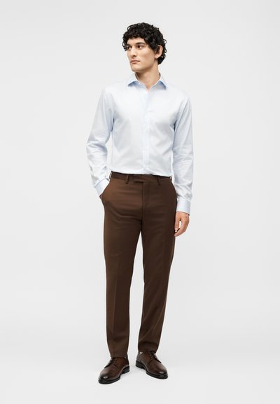 Άνδρας που φοράει ανοιχτό μπλε ριγέ μακρυμάνικο πουκάμισο, καφέ παντελόνι και σκούρα καφέ παπούτσια, στέκεται με το ένα χέρι στην τσέπη.